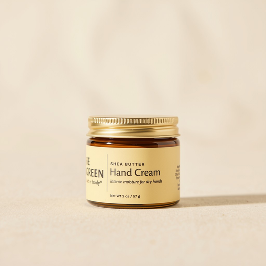 A small amber glass jar of Shea Butter Hand Cream with a gold metal lid and a cream-colored label, set against a warm beige background.