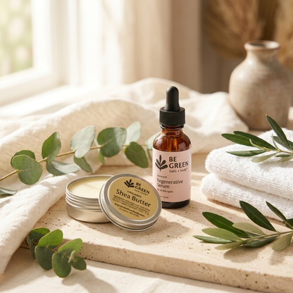 A styled product shot of a small amber dropper bottle labeled BE GREEN Rosehip Facial Serum and a round tin of Be Green Shea Butter, arranged on a stone slab with white towels, eucalyptus leaves, and a soft neutral background.
