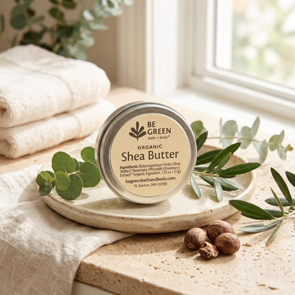 A round tin of BE GREEN Organic Shea Butter sits on a ceramic dish on a stone surface, surrounded by eucalyptus leaves, a nut, and folded towels near a window.