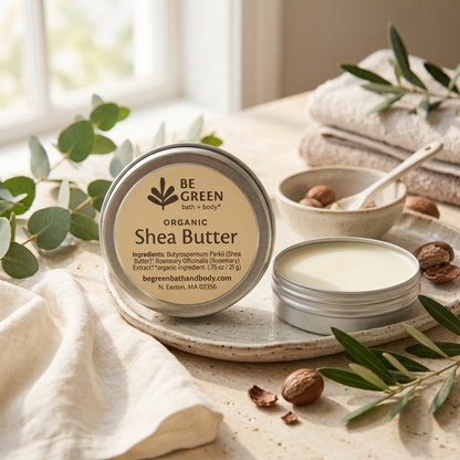 A round tin of BE GREEN Organic Shea Butter sits on a ceramic tray, with the lid placed beside it. The scene is styled with folded beige towels, a small bowl containing shea nuts and a spoon, and green leaves on a light surface near a bright window.