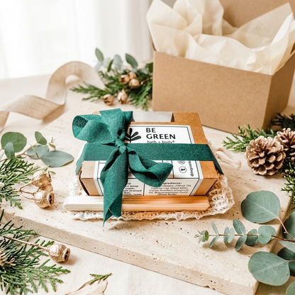 A gift box set from Be Green, featuring a kraft paper box filled with cream tissue paper and a smaller wooden box wrapped in a dark green ribbon, styled with eucalyptus and pinecones on a neutral surface.