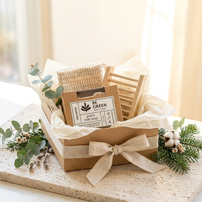 A gift box set containing a brown kraft soap bar labeled 'BE GREEN SOAP CO.' and a small wooden chair-shaped item, arranged with tissue paper, eucalyptus, and cotton stems on a stone surface.