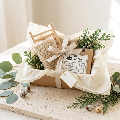 A gift box containing a small wooden chair, a soap bag, and a kraft paper soap box labeled 'BE GREEN goat's milk soap,' wrapped in cream tissue paper and tied with a ribbon, styled with greenery on a stone surface.