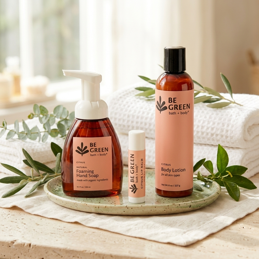 Three BE GREEN bath and body products arranged on a light green tray: a foaming hand soap bottle, a lip balm tube, and a body lotion bottle, styled with white towels and green leaves in a bright bathroom setting.
