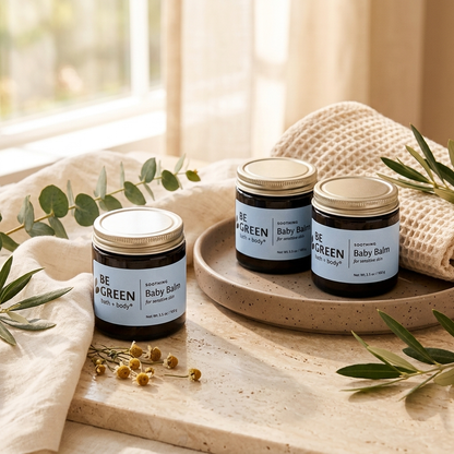 Three jars of BE GREEN Baby Balm arranged on a beige tray and tabletop with linen cloth, eucalyptus leaves, and small flowers in a softly lit indoor setting.