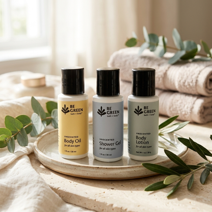 Three BE GREEN bath & body products arranged on a ceramic tray:  Body Oil in yellow,  Shower Gel in pale blue-gray, and Body Lotion in off-white, styled with folded towels, eucalyptus leaves, and soft natural light.