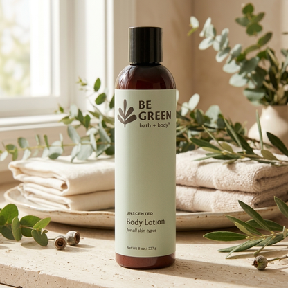 A bottle of BE GREEN bath and body unscented body lotion stands on a stone surface with folded towels, eucalyptus leaves, and a window in the background.