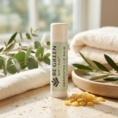 A tube of BE GREEN unscented lip balm stands upright on a stone surface, surrounded by folded cream towels, eucalyptus branches, and small yellow seeds in warm natural light.