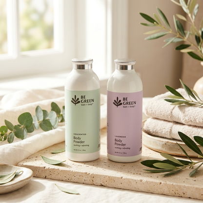 Two bottles of BE GREEN Body Powder standing on a stone tray with folded towels, eucalyptus leaves, and a softly lit window in the background. One bottle has a pale mint label and the other has a pale lavender label.