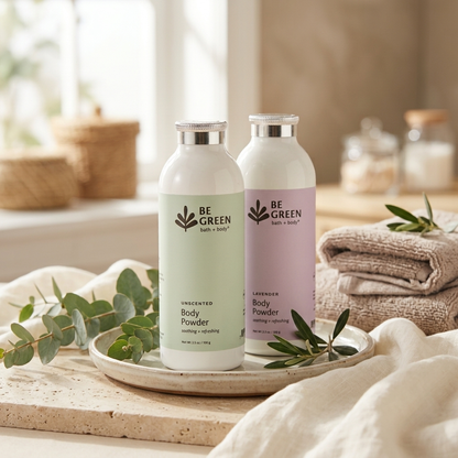 Two BE GREEN body powder bottles on a tray in a softly styled bathroom setting. One bottle is off-white with a pale green label, and the other is off-white with a pale lavender label.