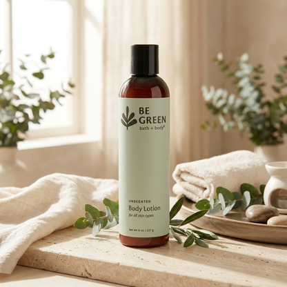 A bottle of BE GREEN Unscented Body Lotion stands on a stone surface in a softly lit bathroom-like setting with folded towels, greenery, and a tray of stones. The product bottle is amber-brown with a pale mint label and a black cap.