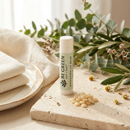 A tube of BE GREEN unscented lip balm stands on a stone slab surrounded by dried flowers, greenery, and folded cream fabric in a softly lit natural setting.
