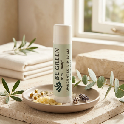 A tube of BE GREEN unscented lip balm stands upright on a small beige dish with dried flowers and greenery, set in a softly lit bathroom-like scene near a window.
