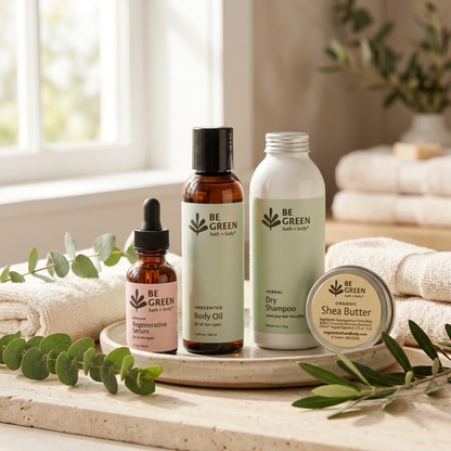 Four BE GREEN bath and body products arranged on a round tray in a bright, softly lit bathroom-like setting with towels, greenery, and a window in the background. The products include a dropper bottle of serum, an amber bottle of unscented body oil, a white bottle of herbal dry shampoo, and a round tin of organic shea butter.
