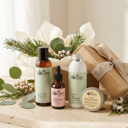 A gift set of BE GREEN natural body care products arranged on a light stone surface with decorative greenery and a wrapped gift box. The set includes a body oil in an amber bottle, a serum in a small amber dropper bottle, a Dry Shampoo in a white bottle, and a shea butter jar, with pale green labels and a kraft gift box tied with a beige ribbon.