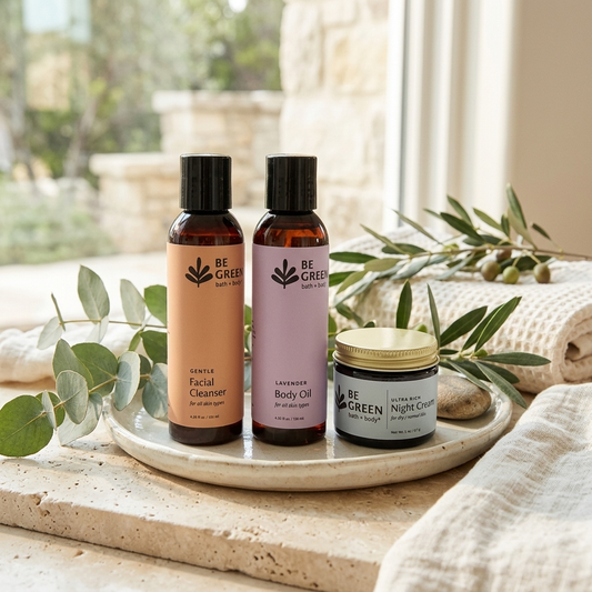 Three BE GREEN skincare products arranged on a round tray: a peach-colored facial cleanser bottle, a lavender body oil bottle, and a black-and-gold night cream jar, styled with eucalyptus leaves and a linen cloth near a window.